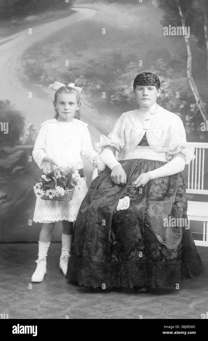 Madre e figlia, 1930 anni, Una ragazza con un vestito bianco che tiene dei fiori, accanto a una donna seduta con un vestito di velluto, foto storica Foto Stock