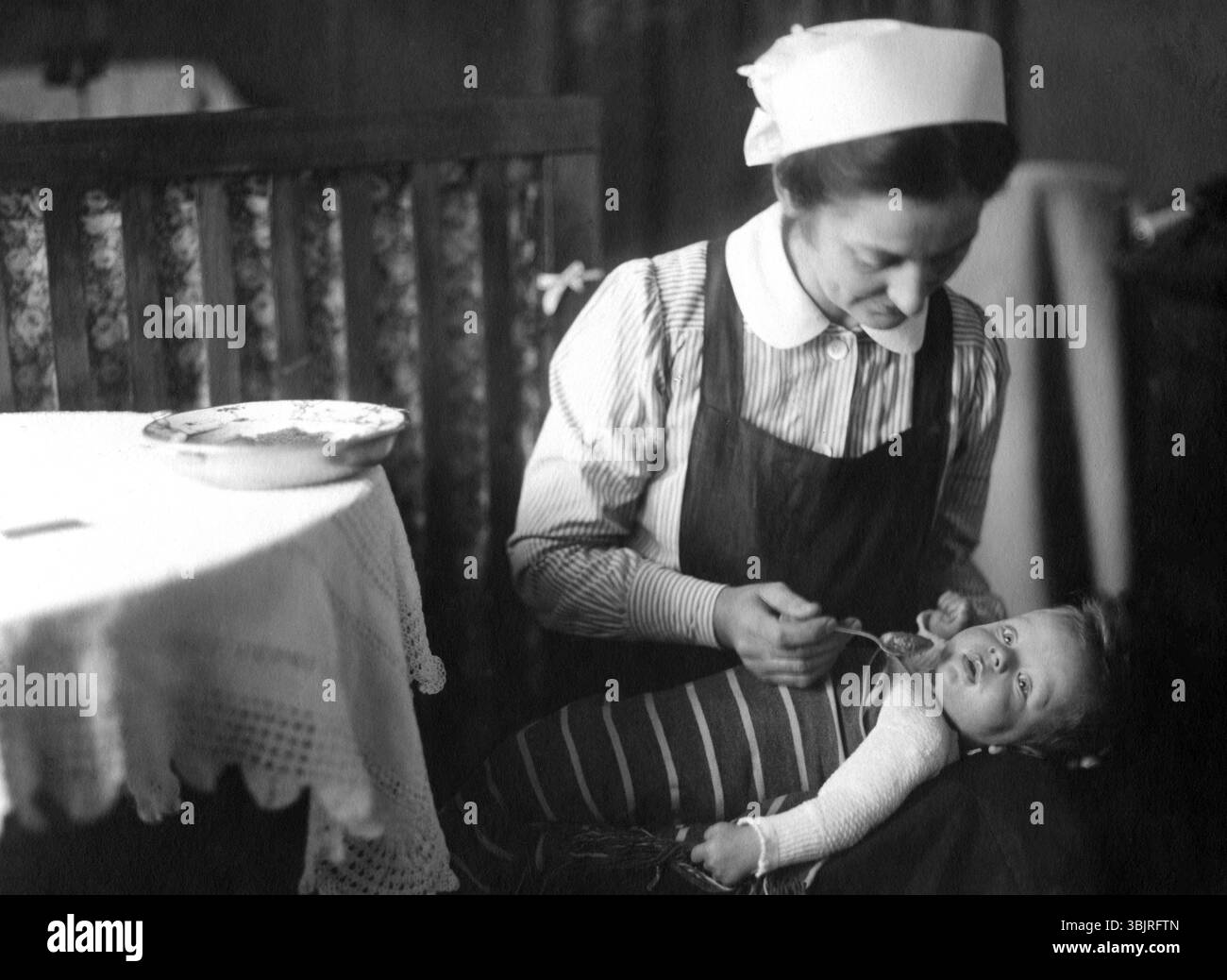 Bambinaia che dà da mangiare a un bambino di 20 anni, 1920 anni, Una donna nutre amorevolmente un bambino in un ambiente storico domestico, mostrando una foto attenta e storica Foto Stock