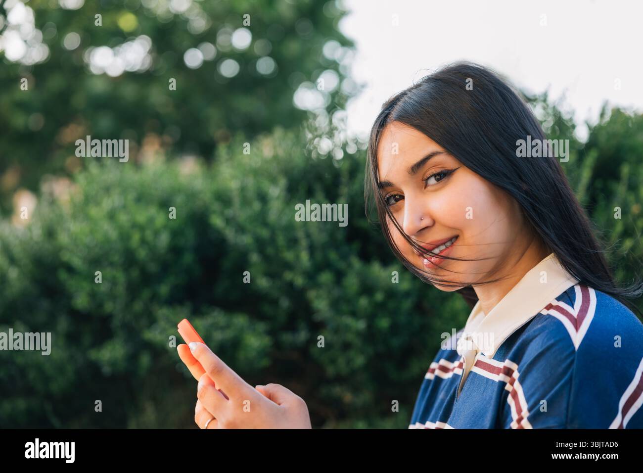 Una giovane donna con una polo a righe sorride davanti alla fotocamera mentre tiene in mano uno smartphone, in piedi di fronte a una vegetazione lussureggiante con i capelli che soffiano delicatamente Foto Stock