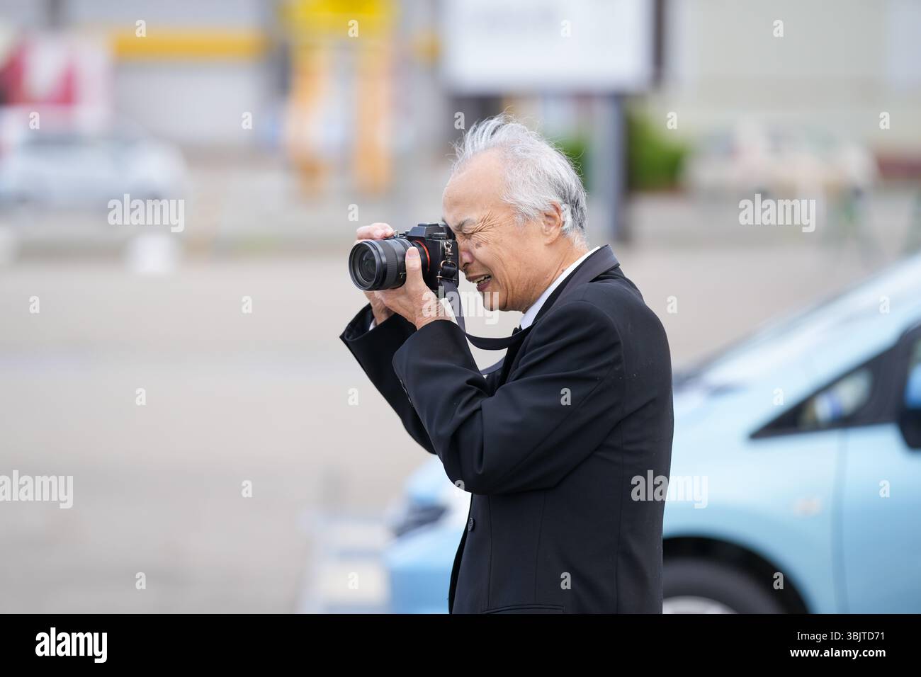In un parcheggio giapponese, un uomo giapponese alla fine degli anni '70 indossa una tuta nera e tiene in mano una macchina fotografica mirrorless, inquadrando accuratamente uno scatto con occhi a fuoco Foto Stock