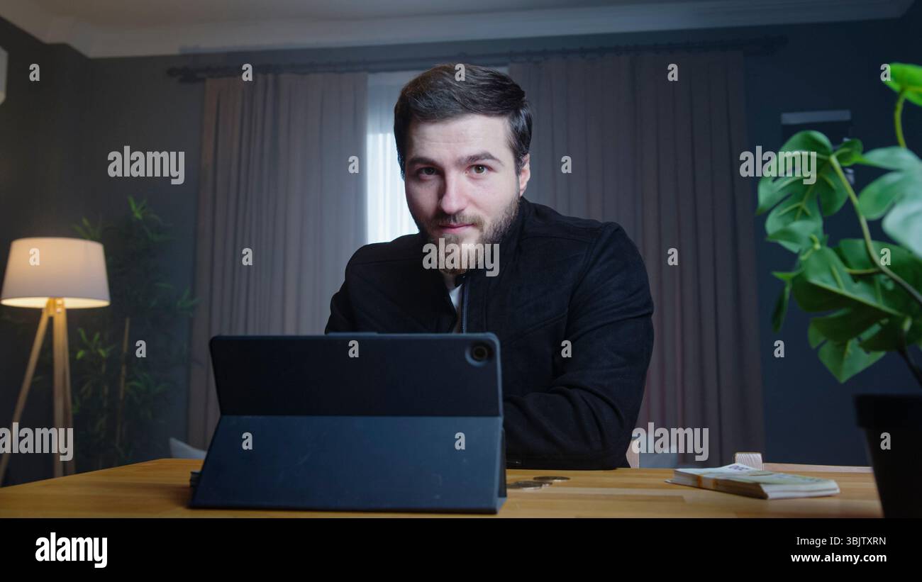 Un giovane con una giacca nera guarda la fotocamera mentre è seduto alla scrivania con un tablet, suggerendo sicurezza e comunicazione digitale. Foto Stock