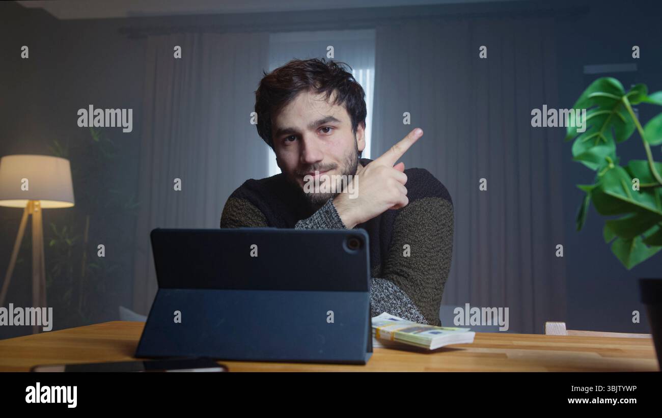 Un giovane uomo si siede alla scrivania utilizzando un tablet, puntando verso l'alto con i soldi sul tavolo, suggerendo business online o successo. Foto Stock