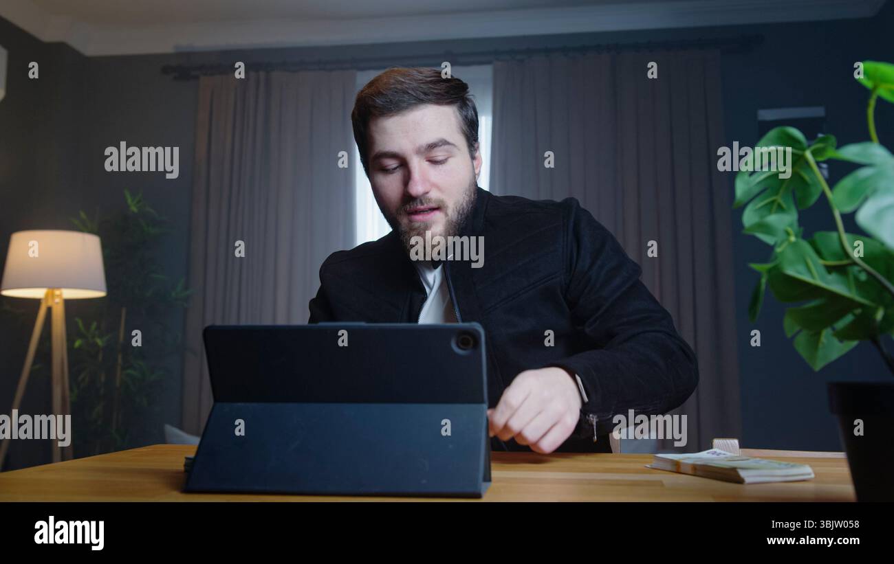 L'uomo con barba in giacca nera si siede alla scrivania utilizzando un tablet, concentrato e premuroso, con una pila di contanti nelle vicinanze di un ufficio domestico. Foto Stock