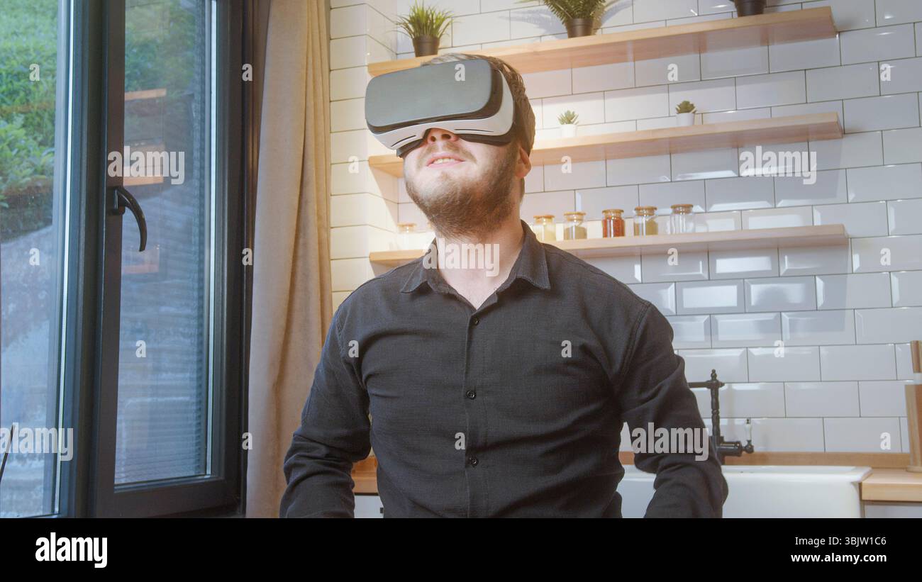 L'uomo barbuto in camicia nera utilizza un visore VR in una cucina moderna e luminosa, godendo di coinvolgenti contenuti digitali o esplorazioni virtuali. Foto Stock