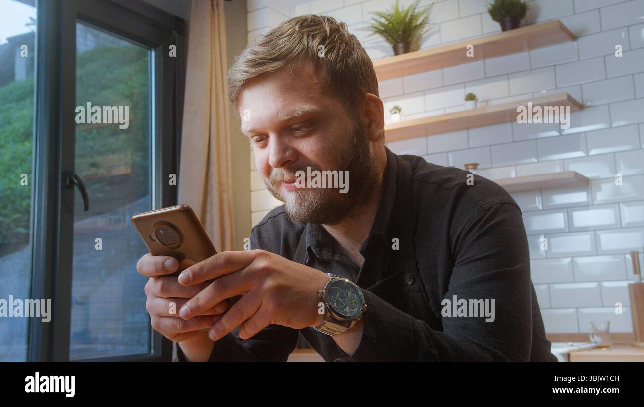 L'uomo barbuto con una camicia nera sorride mentre guarda il suo smartphone, magari guardando un messaggio o navigando in un ambiente accogliente. Foto Stock