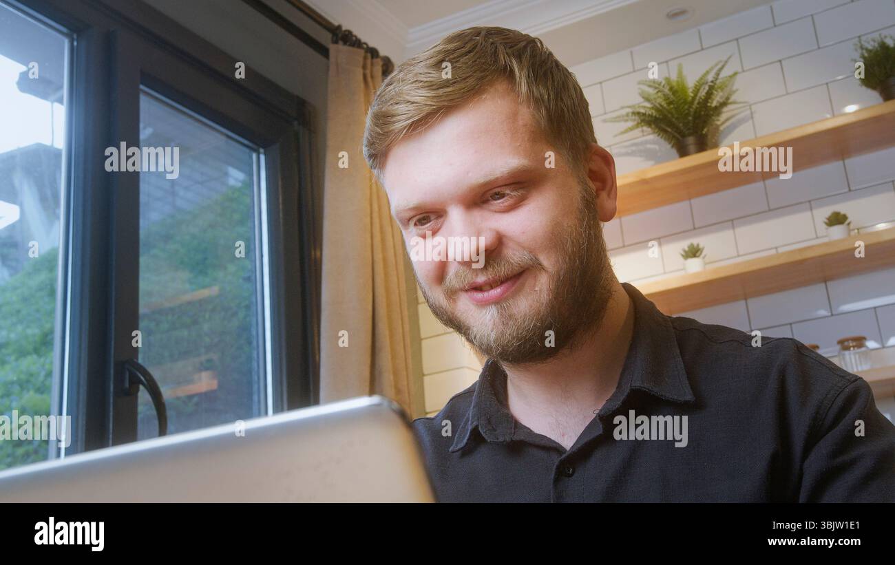 Uomo allegro sorridente mentre guarda lo schermo del laptop vicino alla finestra in un'accogliente cucina, che suggerisce buone notizie o una piacevole attività online. Foto Stock