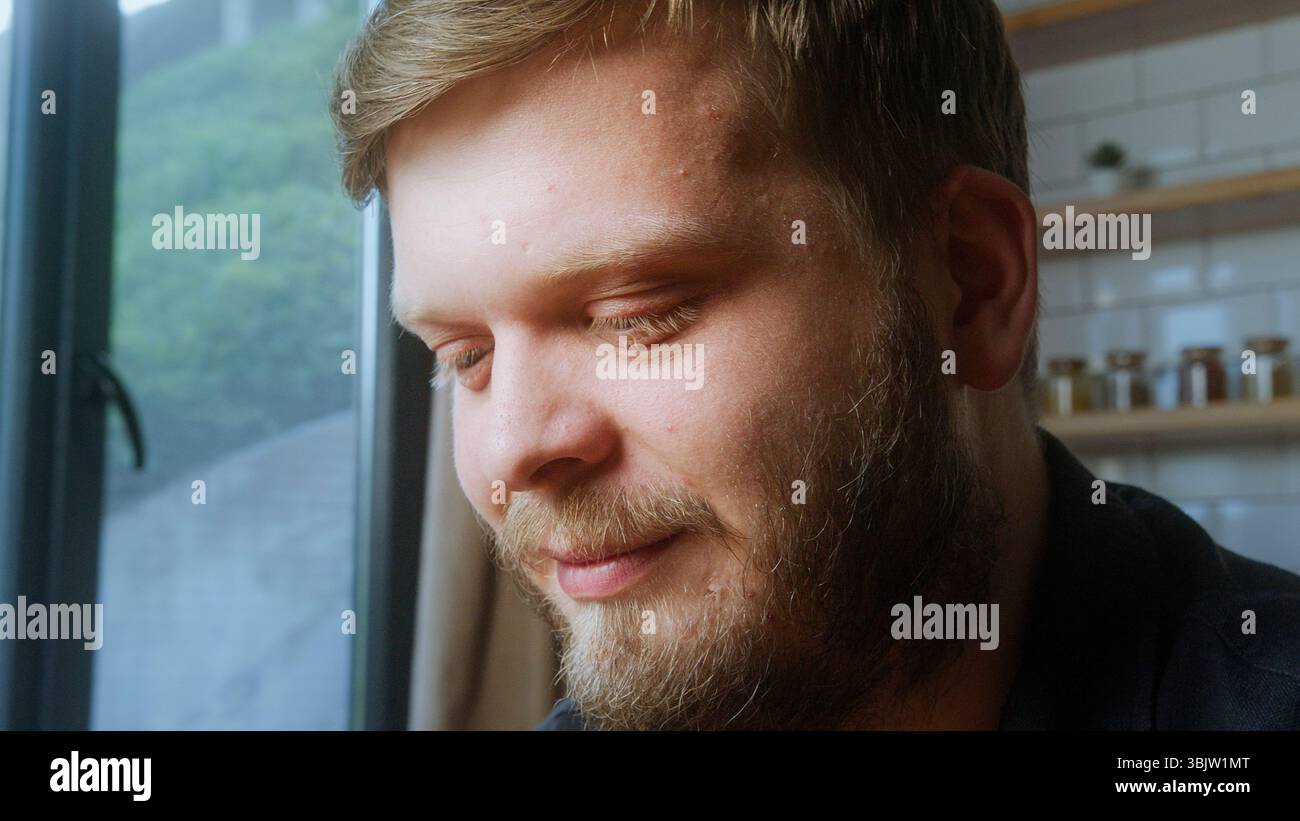 L'uomo barbuto con un sorriso gentile guarda verso il basso vicino a una finestra, catturata dalla luce naturale soffusa, con un momento tranquillo e riflessivo. Foto Stock