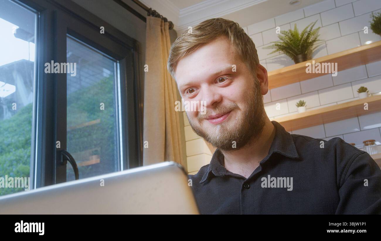 L'uomo barbuto sorride con gioia mentre guarda uno schermo di un notebook vicino a una finestra, suggerendo comunicazioni online o notizie positive. Foto Stock