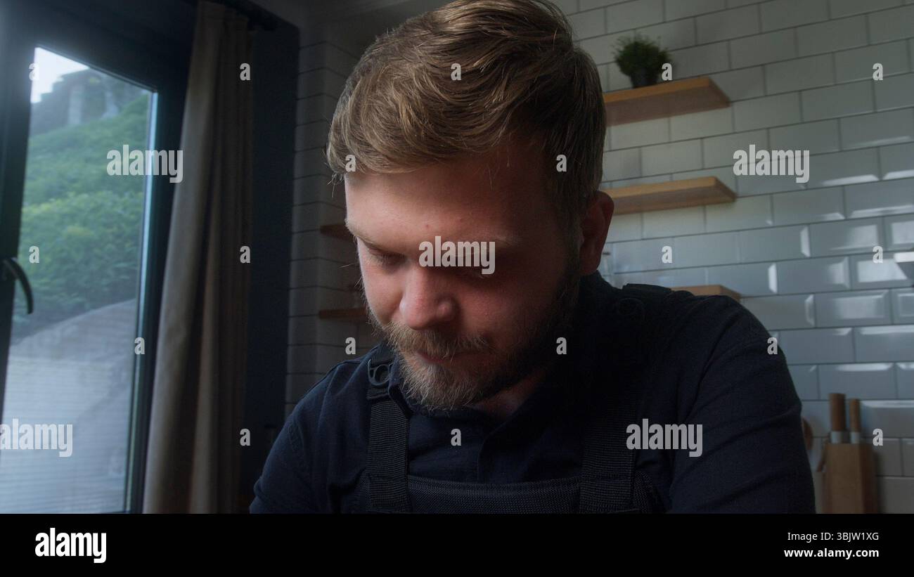 L'uomo barbuto guarda in basso con un'espressione seria in una stanza delicatamente illuminata, suggerendo un pensiero profondo, una riflessione o una preoccupazione. Foto Stock