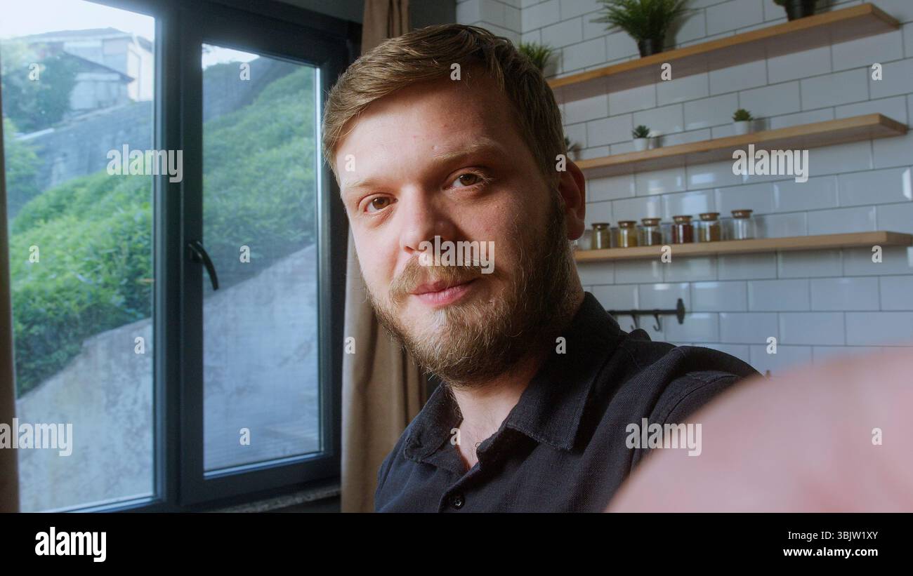 L'uomo barbuto guarda direttamente la fotocamera con un sorriso dolce, suggerendo un momento di amicizia durante una videochiamata in piena luce naturale. Foto Stock