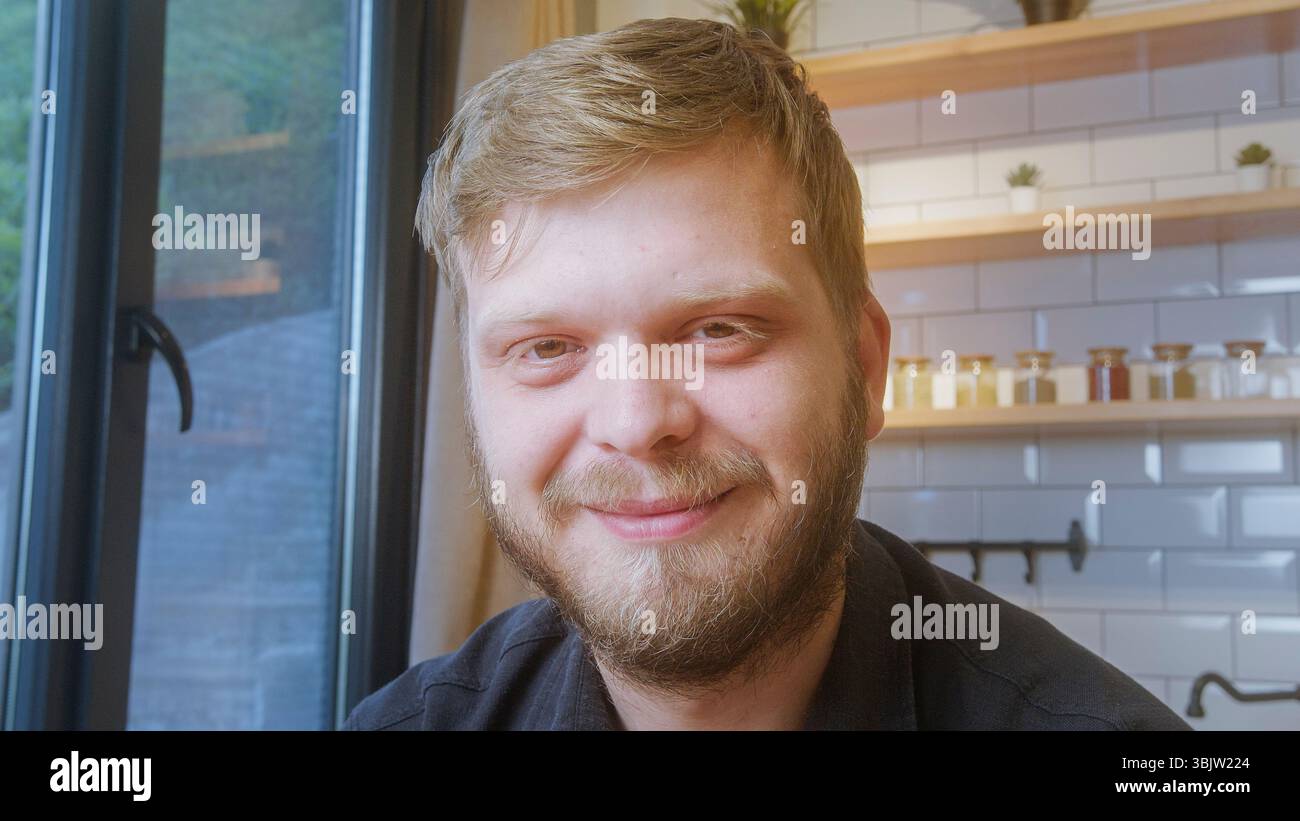 Primo piano di un uomo allegro e barbuto che sorride calorosamente mentre si siede vicino a una finestra in uno spazio interno accogliente e ben illuminato. Foto Stock