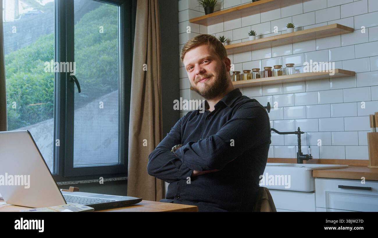 L'uomo barbuto in camicia nera si stende con le braccia incrociate, sorridendo con sicurezza vicino a una finestra in un'accogliente cucina o in un ambiente domestico. Foto Stock