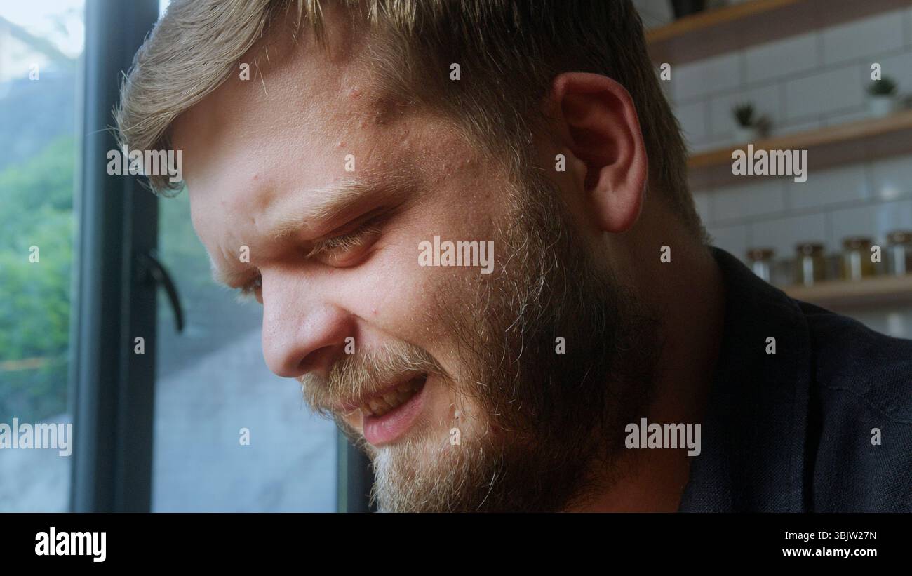 L'uomo barbuto vicino a una finestra mostra un'espressione emotiva, che potrebbe indicare dolore, angoscia o riflessi profondi nella luce naturale. Foto Stock