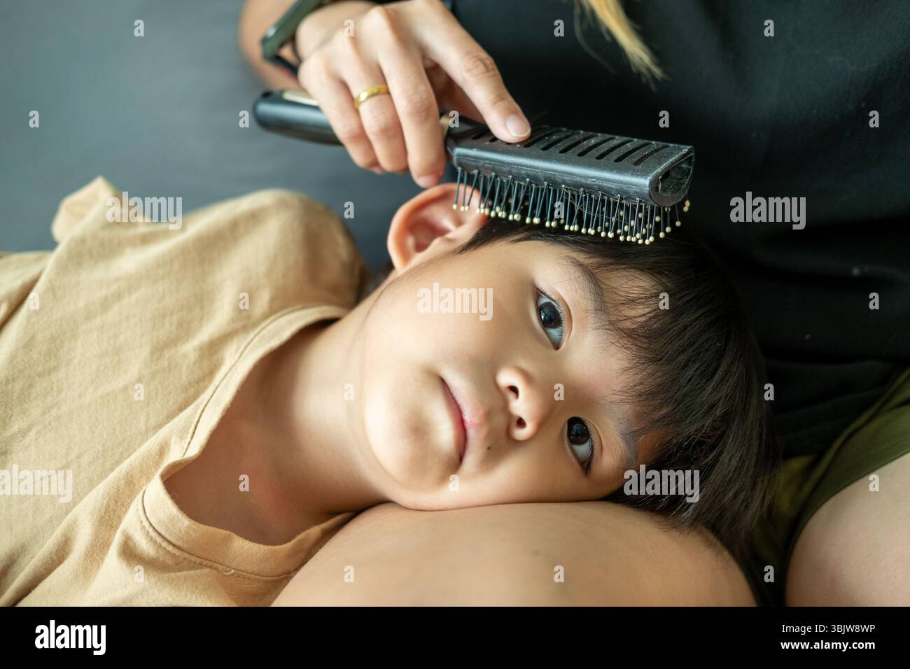 Il ragazzo asiatico giace in grembo di sua madre, usando un pettine per pettinarle i capelli. Foto Stock