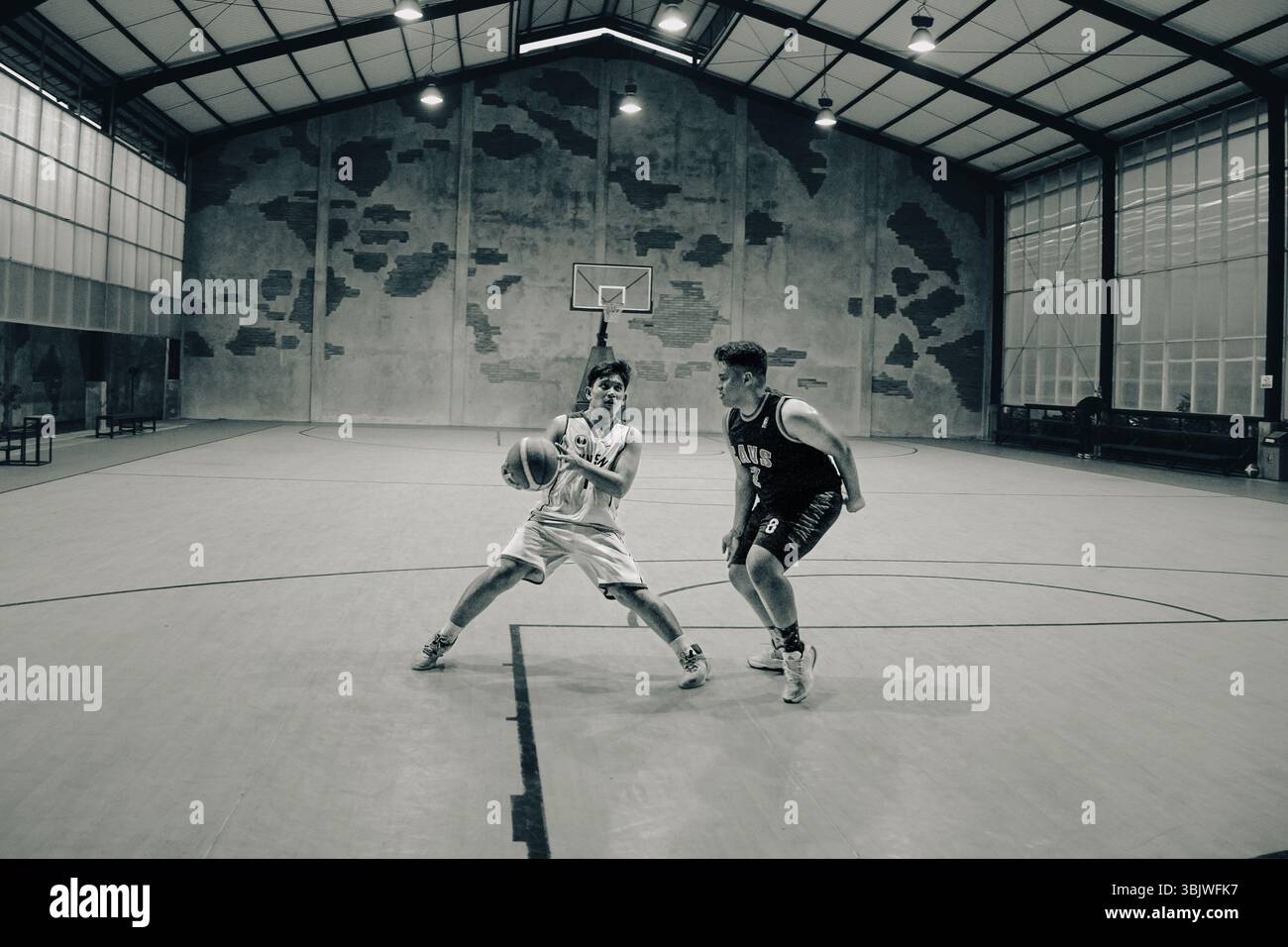 In un grintoso campo al coperto in stile magazzino, due giovani uomini sono bloccati in un'intensa partita di basket uno contro uno Foto Stock