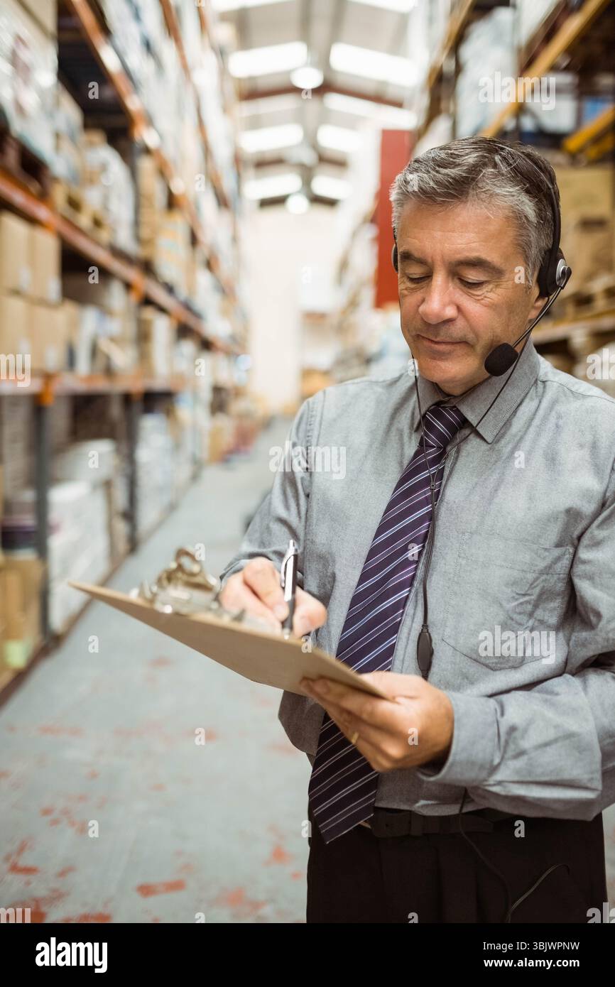 Uomo che supervisiona l'inventario nella corsia del magazzino indossando cravatta, tenendo appunti e cuffie, spazio per le copie Foto Stock