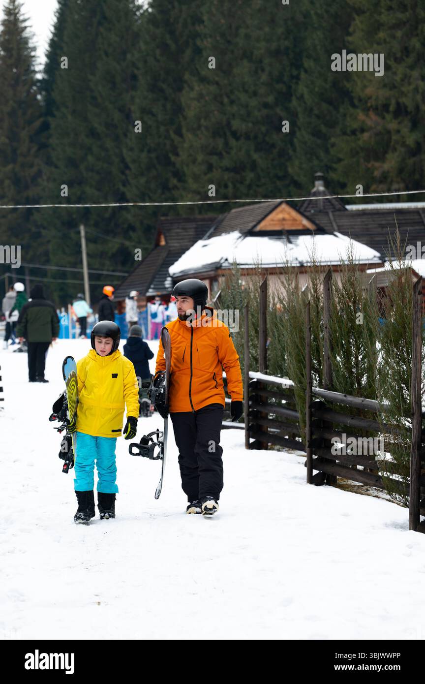 Due bambini passeggiano attraverso un paesaggio innevato vicino a un'accogliente stazione sciistica, portando con sé l'attrezzatura da sci e godendosi l'allegra atmosfera degli sport invernali. Foto Stock