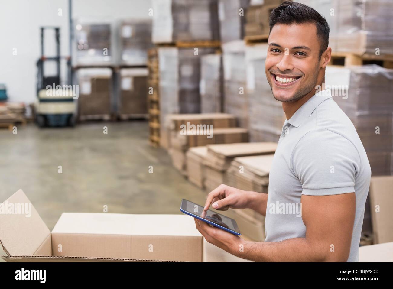 Lavoratore di sesso maschile che indossa una polo grigia chiaro che scansiona l'inventario su un tablet accanto alla scatola aperta in magazzino Foto Stock