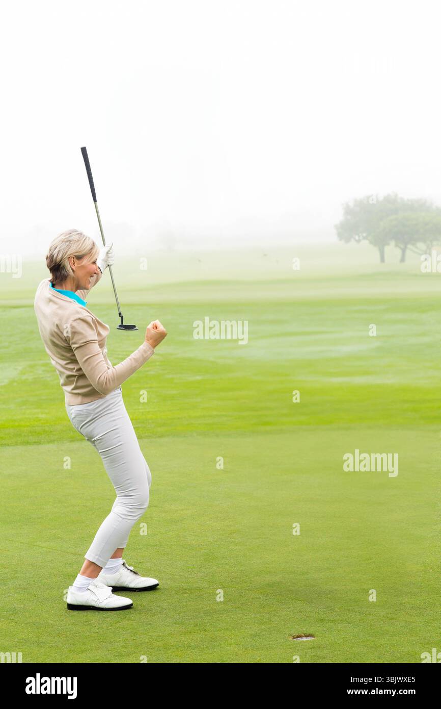 Golfista donna anziana che celebra il putt affondato su un putting green nebbioso con pugno e putter con guanti Foto Stock