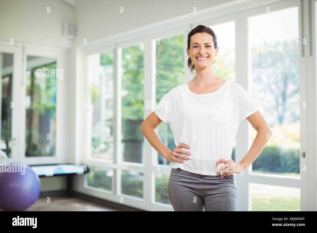 Donna in piedi accanto alla palla di stabilità viola e alla panchina nella sala fitness accanto a grandi finestre, spazio fotocopie Foto Stock