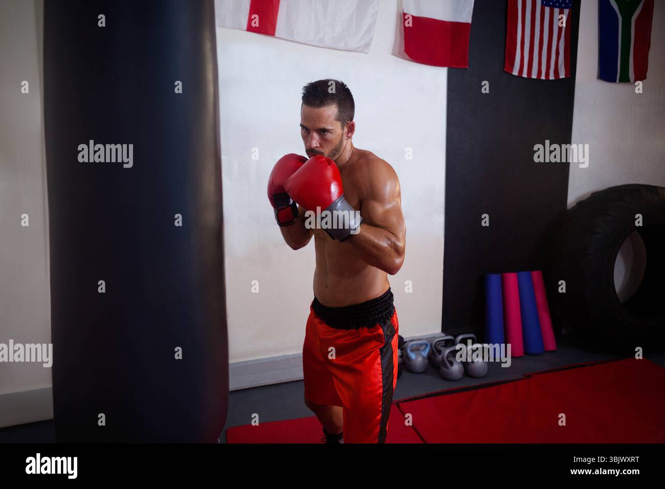 Pugile maschile in piedi in guardia nella palestra di boxe con guanti da boxe rossi e pantaloncini di fronte al sacchetto di punzonatura Foto Stock