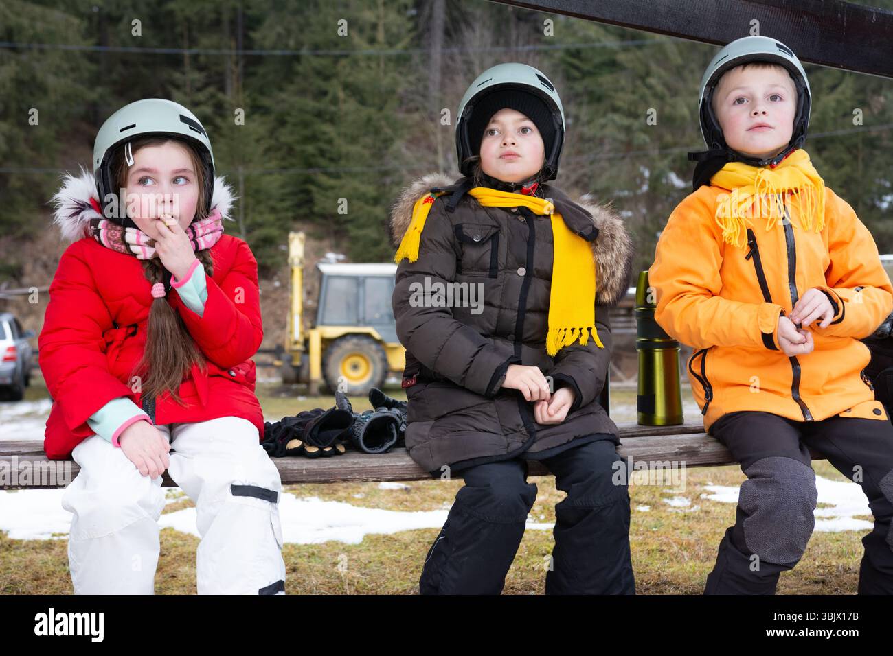 Tre bambini seduti insieme su una panchina, vestiti caldi per le attività invernali. I loro volti ansiosi suggeriscono il divertimento e l'emozione di sciare o snowboardi Foto Stock