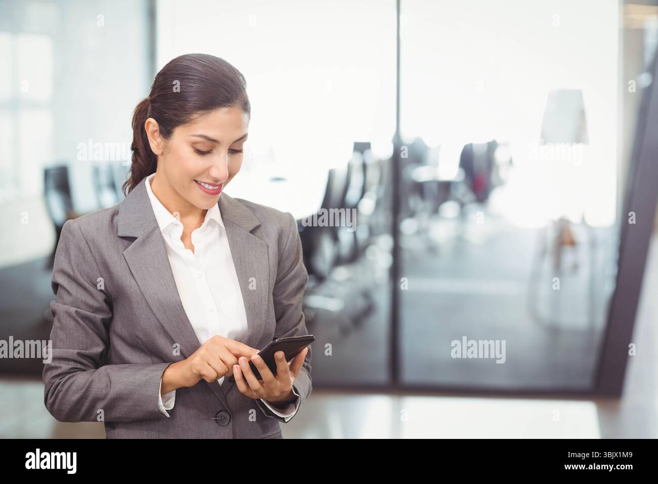Donna asiatica che indossa una tuta grigia che utilizza lo smartphone in ufficio tramite partizione di vetro, spazio copia Foto Stock