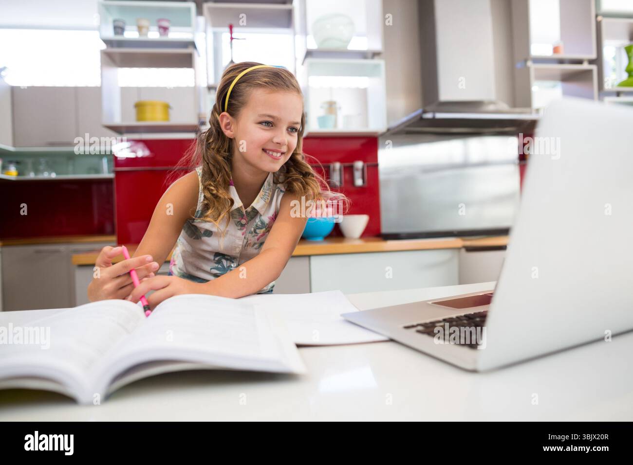 Studentessa in età scolare che studia i compiti al bancone della cucina con laptop, libro di testo, penna e ciotole da mescolare Foto Stock