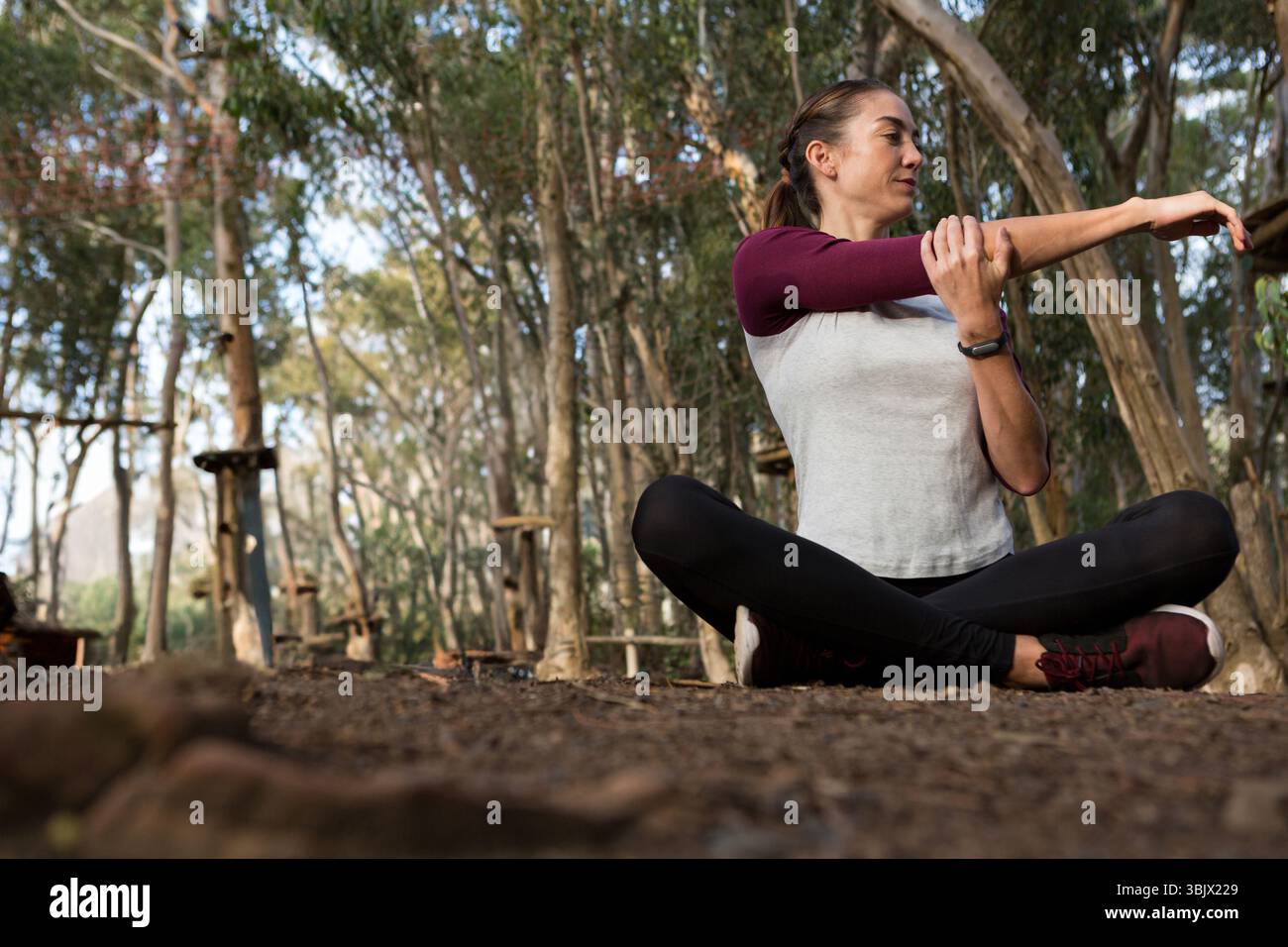 Donna che si allunga il braccio destro sul petto in un percorso di corda forestale indossando orologi, scarpe da ginnastica e spazio per copiare Foto Stock