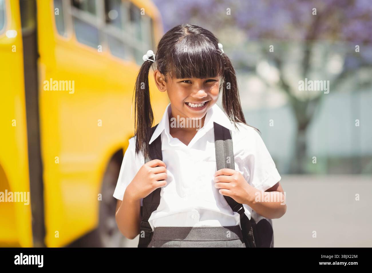 Bambina in uniforme scolastica in piedi sul marciapiede di scuolabus giallo, con zaino nero Foto Stock