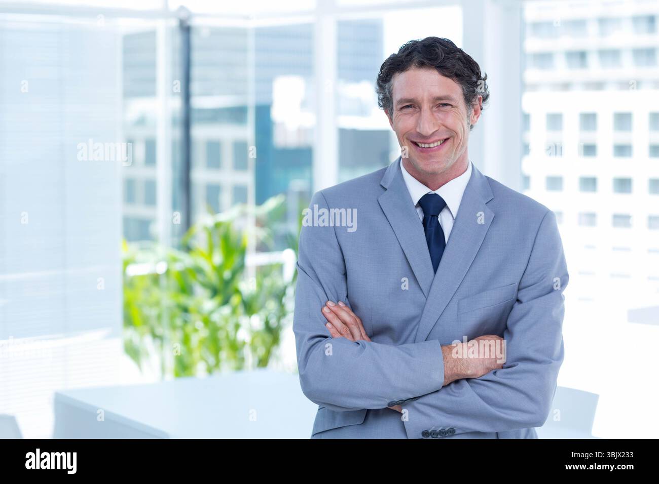 Uomo d'affari che indossa una tuta grigia e una cravatta blu navy, in piedi in ufficio con la pianta della scrivania vicino alle finestre dello skyline Foto Stock