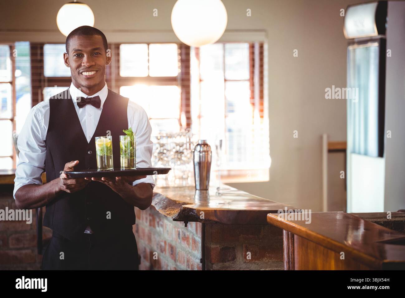 Uomo afroamericano in abito formale che serve cocktail su vassoio nero al rustico bar, spazio per fotocopie Foto Stock