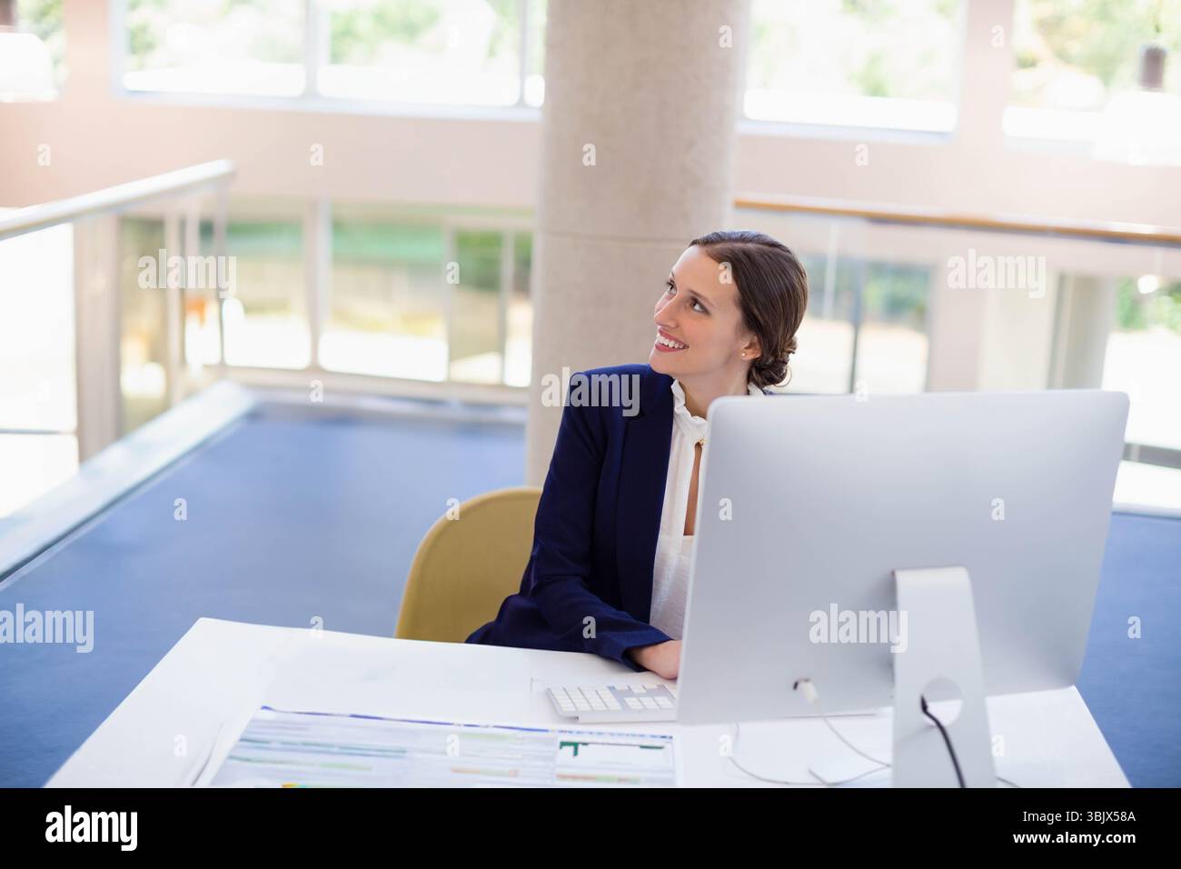 Lavoratrice che indossa un blazer blu scuro sorridente, guarda il monitor alla scrivania dell'ufficio, copia spazio Foto Stock