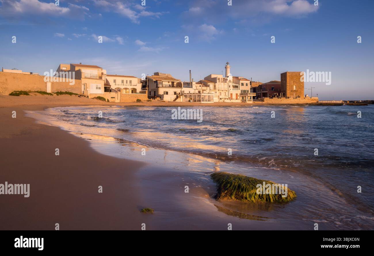 Le onde si infrangono sulla riva mentre il tramonto bagna gli edifici e la spiaggia di Punta secca in un caldo bagliore, Sicilia, Italia Foto Stock