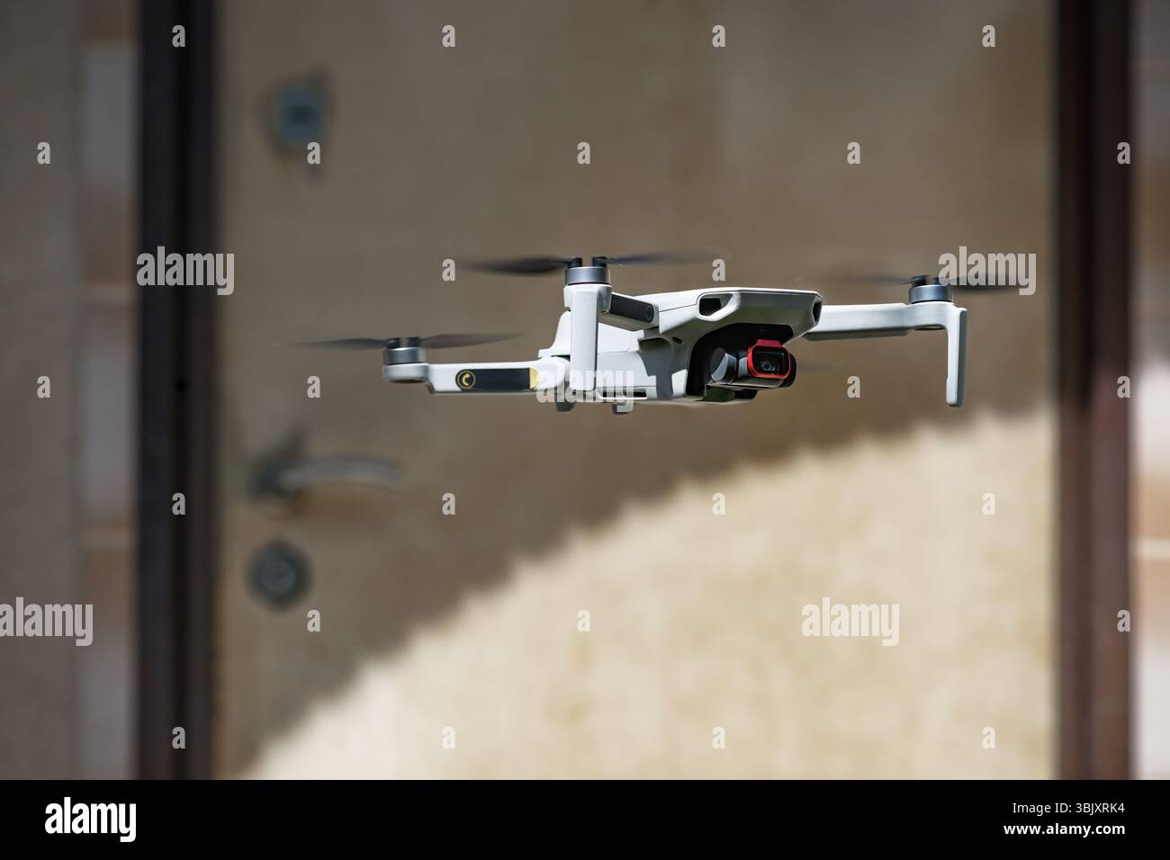 Piccolo drone volante Quadcopter in modalità volo con fotocamera all'aperto di fronte alla porta di un edificio residenziale sullo sfondo ombreggiato Foto Stock