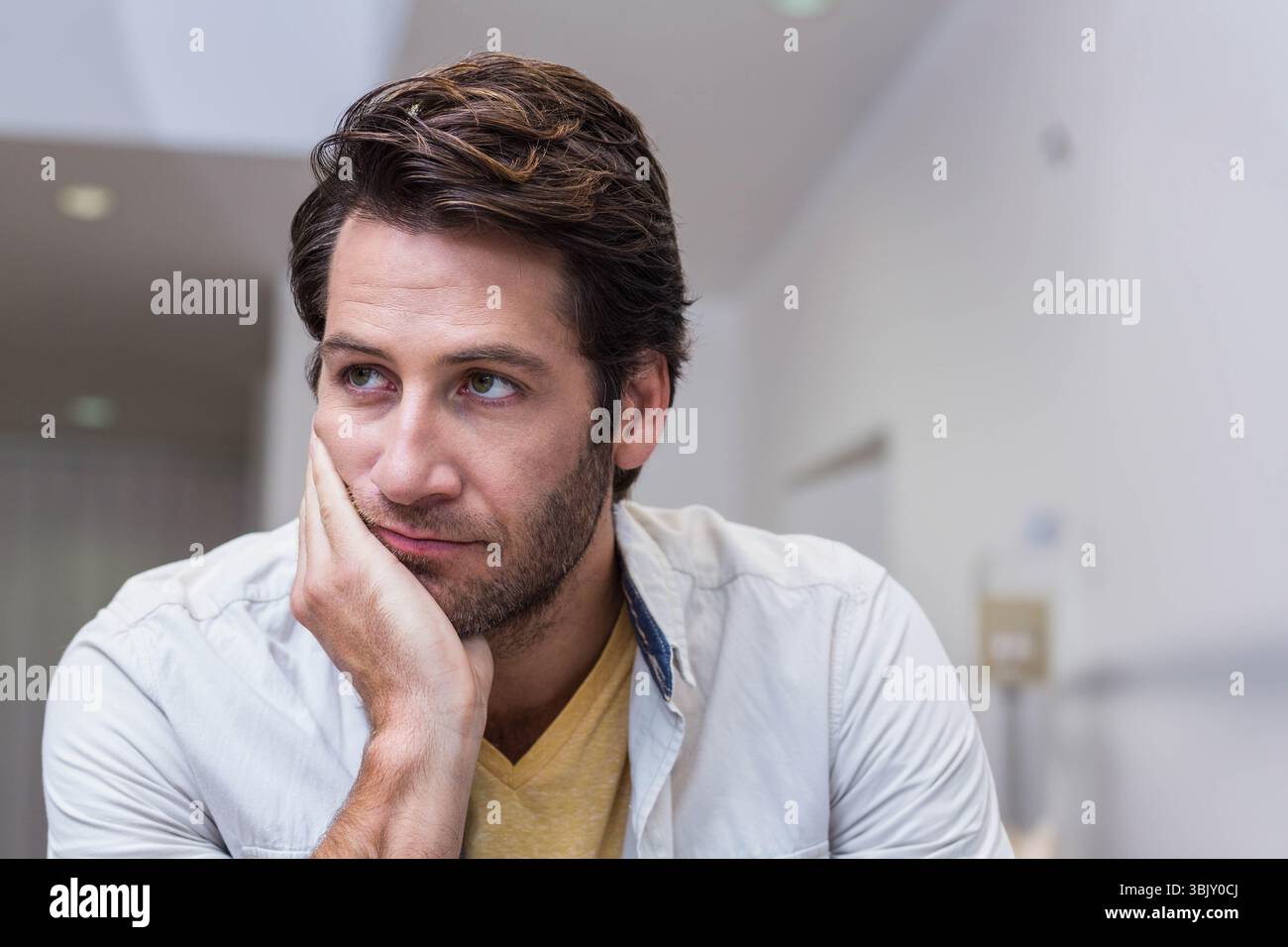 Uomo che poggia il mento sulla sinistra, che guarda a destra vicino a un pannello montato a parete sotto le luci incassate in ufficio Foto Stock