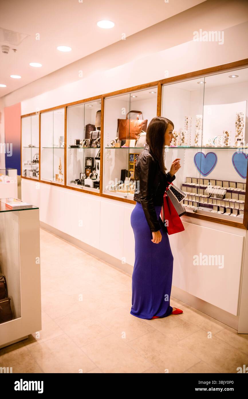 Donna che indossa una giacca in pelle accanto all'esposizione di gioielli in vetro, tenendo in mano borse rosse e a righe Foto Stock