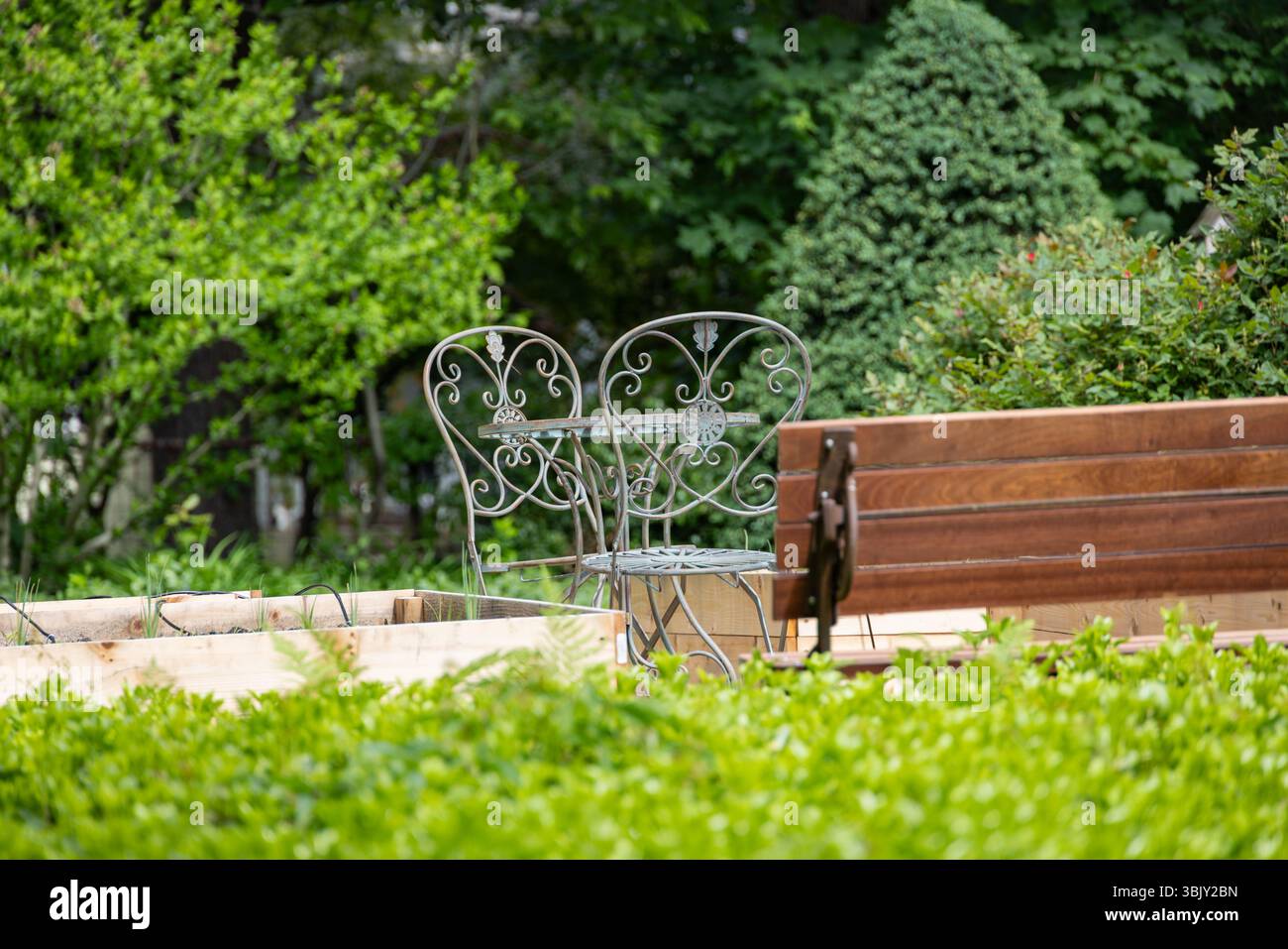 Tavolo decorativo da giardino in ferro con sedie e panca in legno nel verde lussureggiante, tranquillo ambiente esterno per riposare, conversare o rilassarsi nella natura Foto Stock