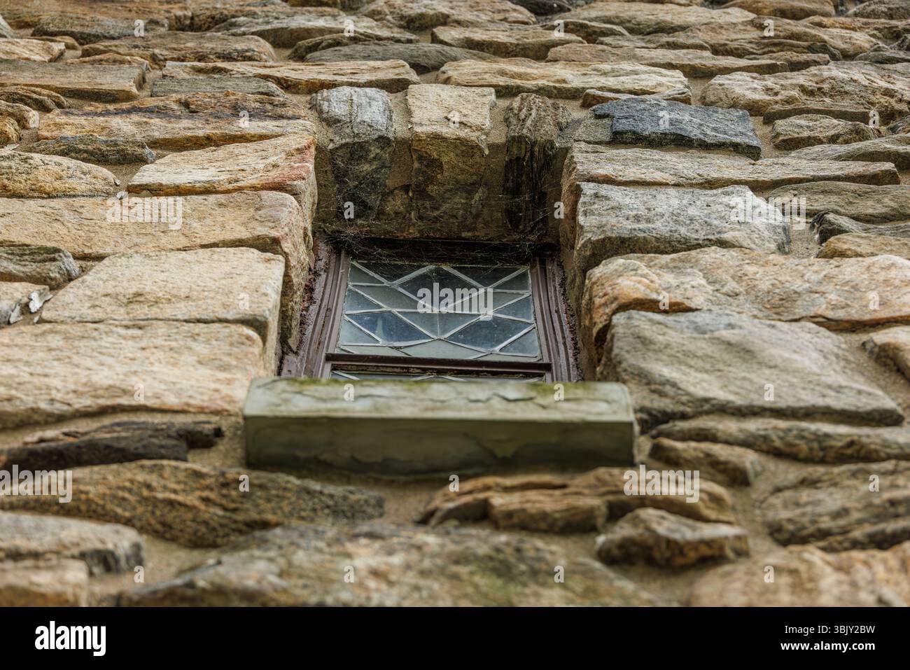 Foto a colori di una piccola finestra geometrica su un muro di pietra, vista dal basso, che mostra una trama ruvida, muratura storica e dettagli architettonici tradizionali. Foto Stock