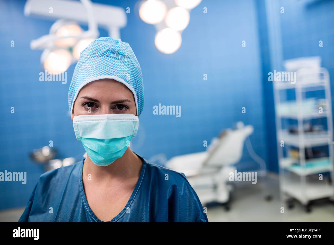Chirurgo donna che indossa scrub, cappuccio e maschera sotto le luci chirurgiche in sala operatoria con carrello Foto Stock
