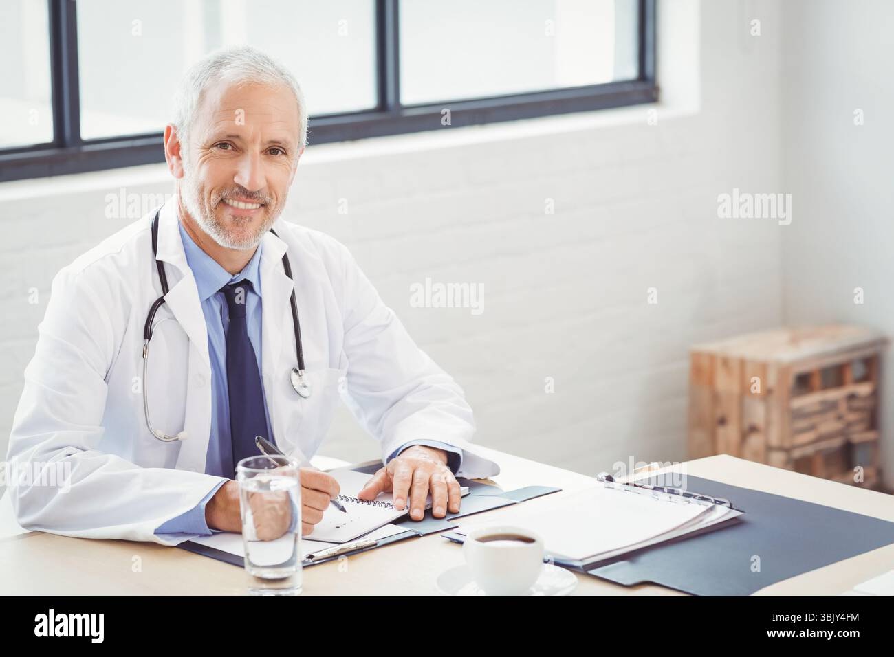 Il medico senior scrive appunti alla reception nella sala consultiva con stetoscopio, tazza da caffè e appunti Foto Stock