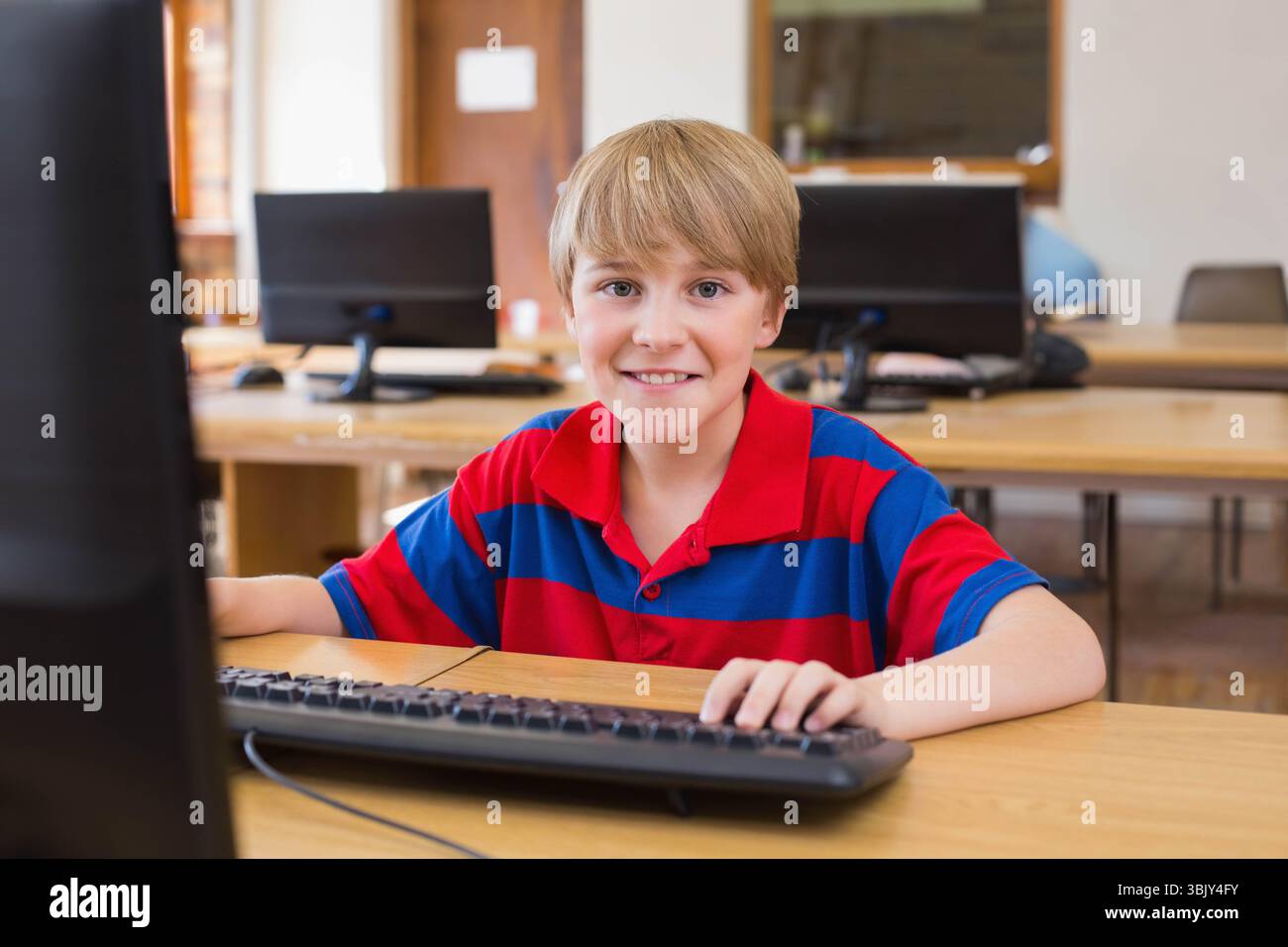 Ragazzo preadolescente seduto alla scrivania di legno nel laboratorio di computer della scuola usando il computer desktop e il mouse Foto Stock