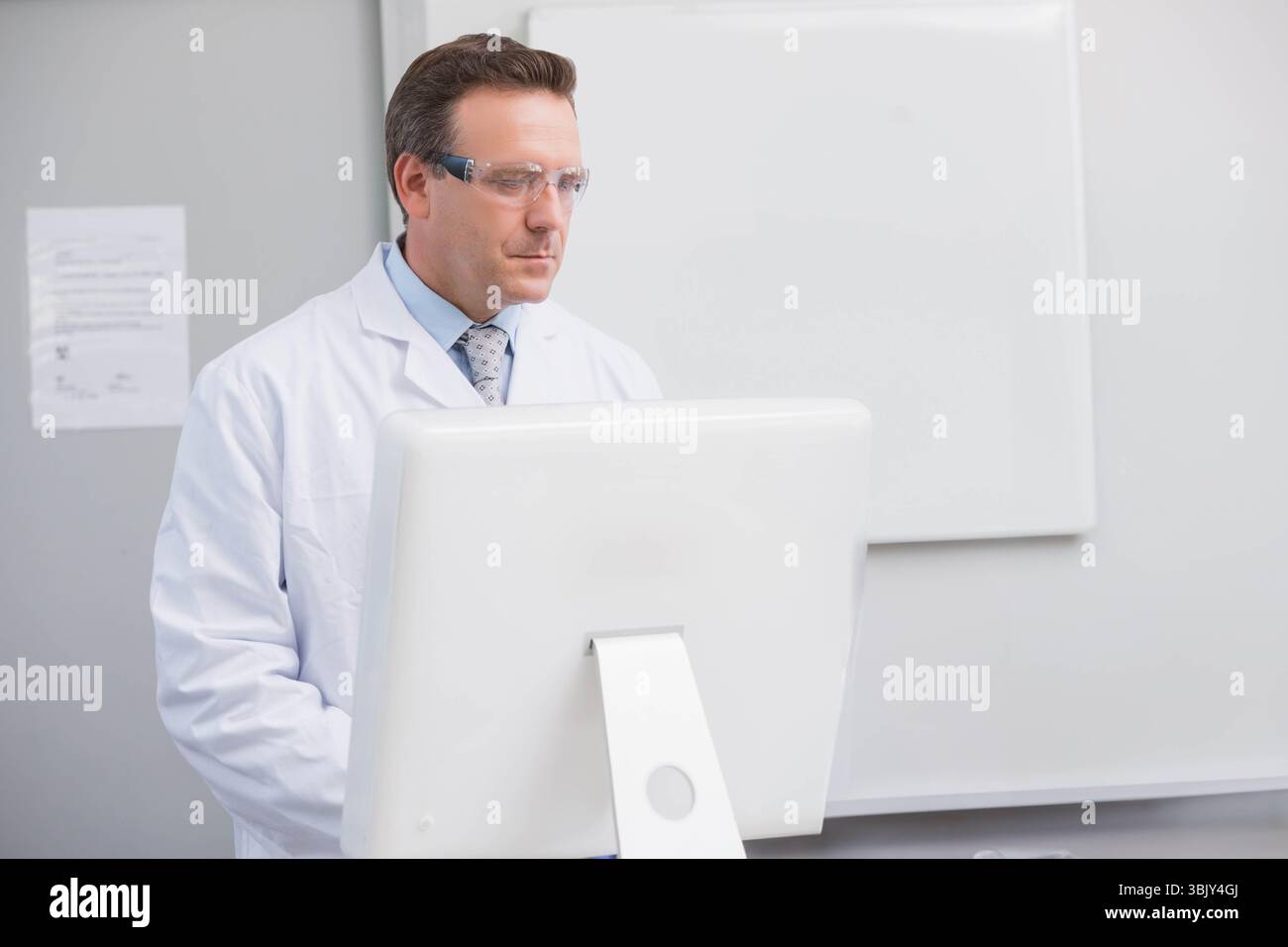 Scienziato maschio che indossa camice da laboratorio e occhiali di sicurezza, che esamina il monitor del computer in laboratorio, copia spazio Foto Stock