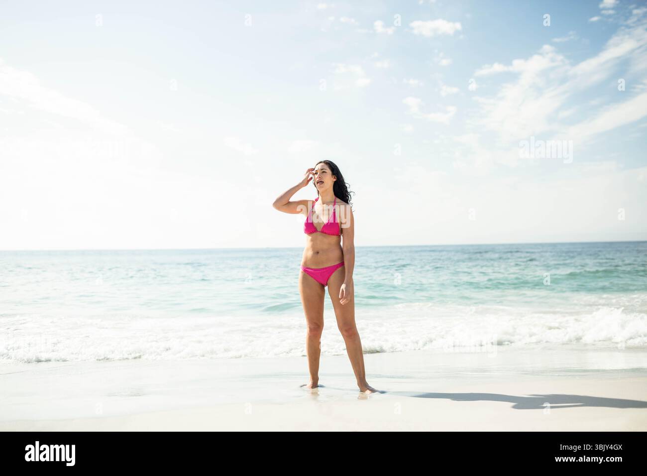 Donna a piedi nudi sul bordo della spiaggia che indossa un bikini rosa, che protegge gli occhi e guarda verso l'orizzonte Foto Stock