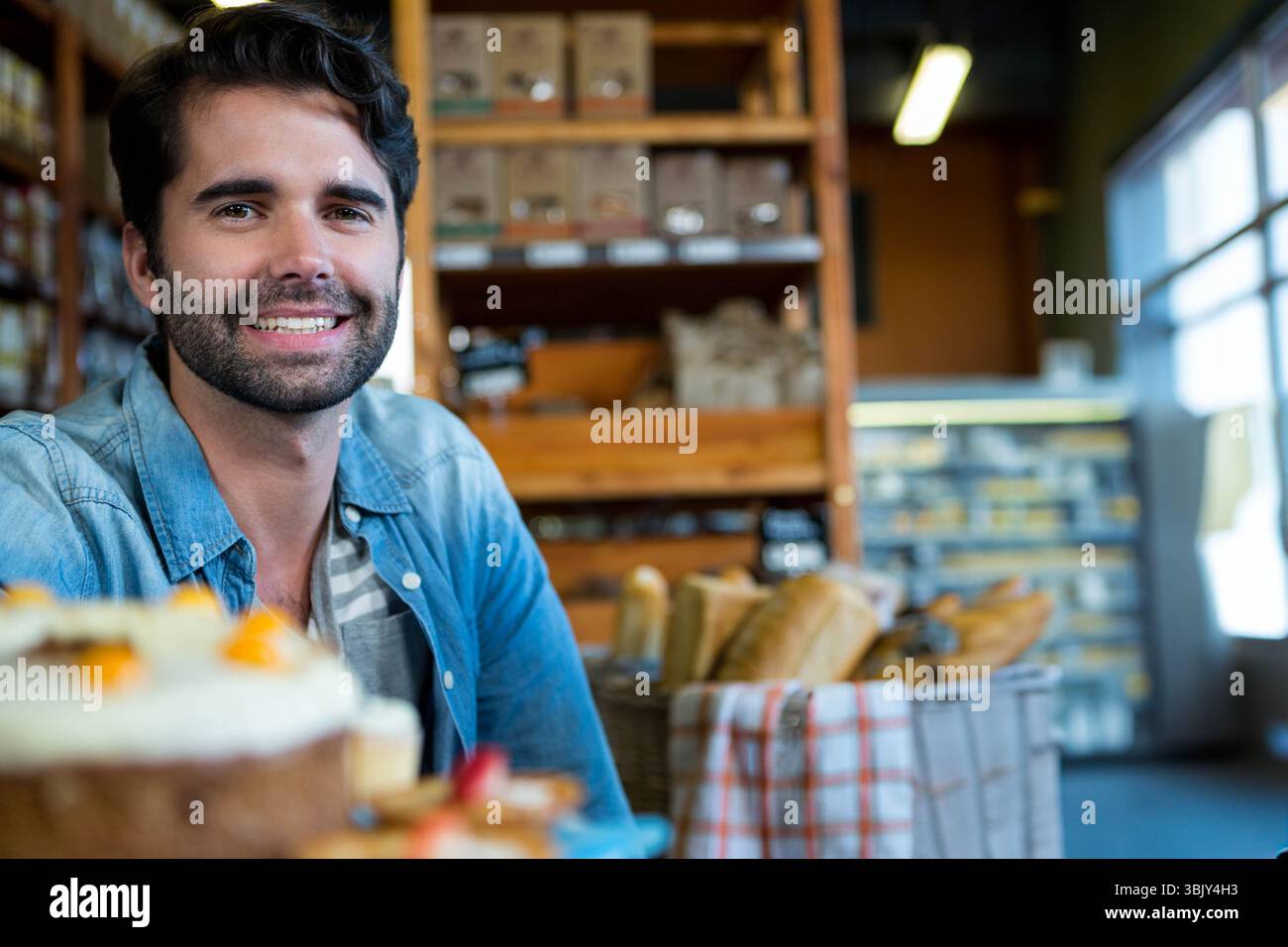 Uomo sorridente che indossa una camicia in denim assaggia una torta con contorno di frutta al tavolo del panificio vicino ai cesti del pane Foto Stock