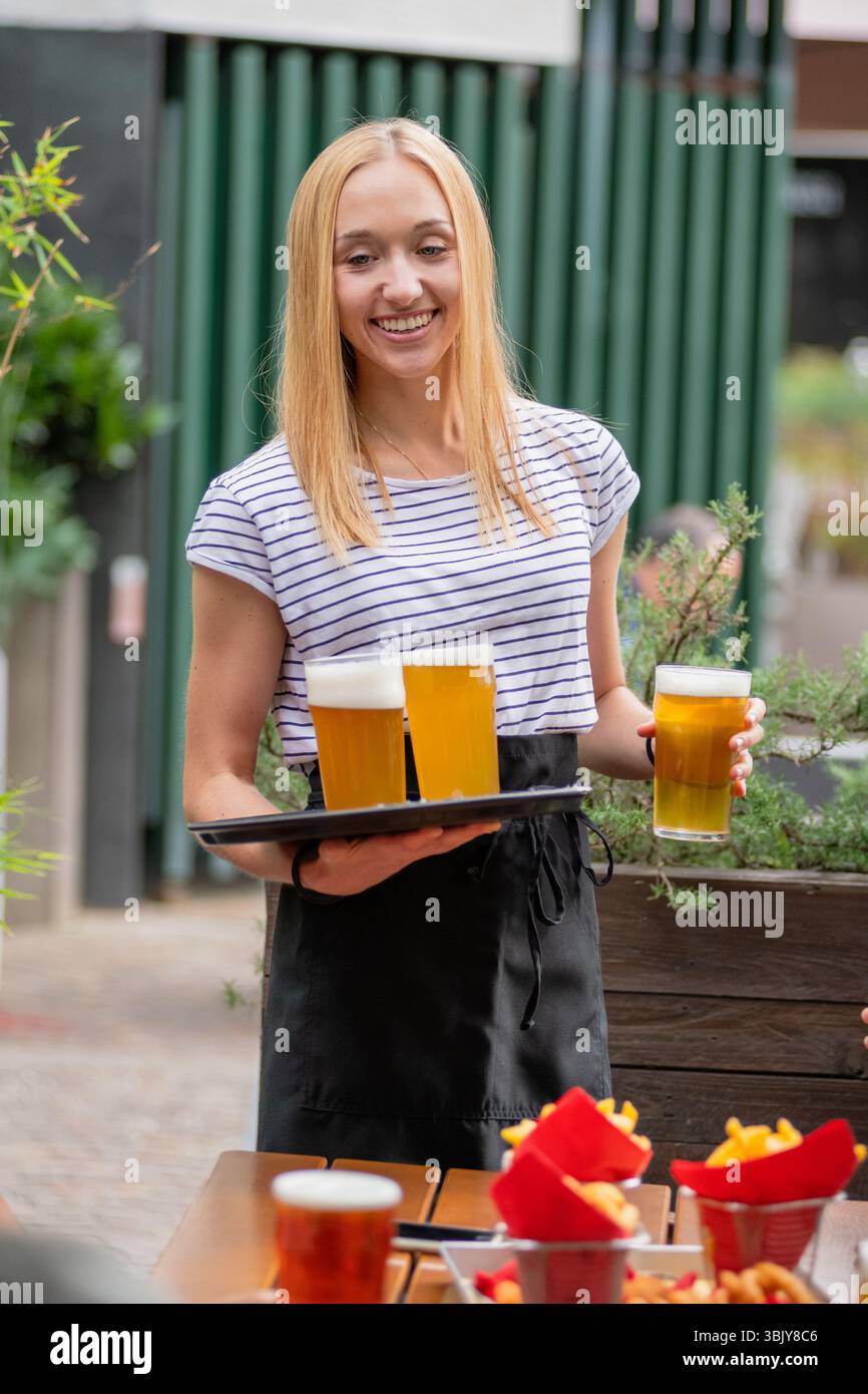 Cameriera sorridente che porta con sé un vassoio di birre al servizio dei clienti presso il ristorante all'aperto Foto Stock