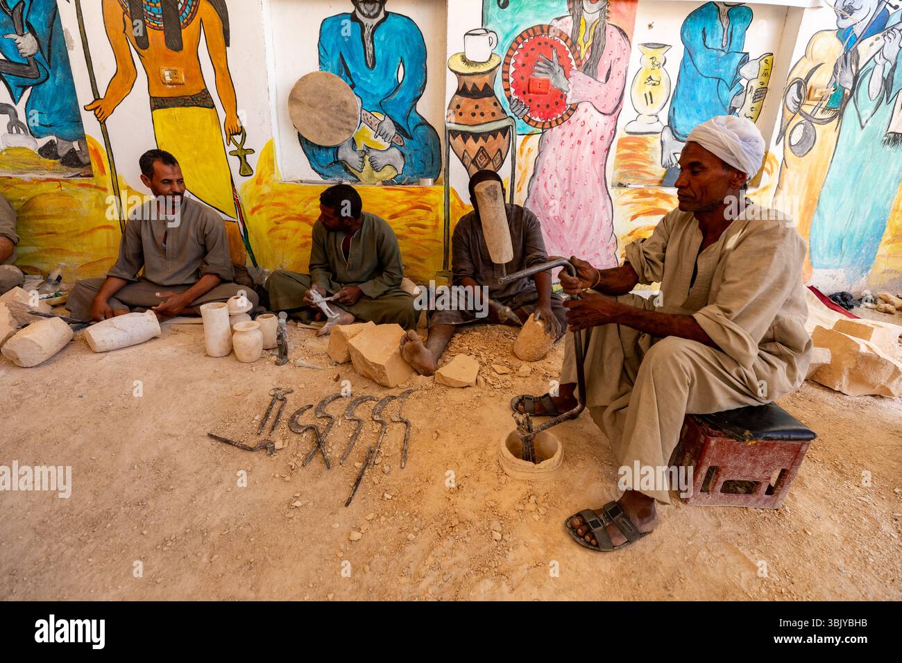 In questa immagine, un gruppo di artigiani egiziani è mostrato mentre realizzano manufatti in pietra, probabilmente in vendita ai turisti. L'ambiente sembra essere un laboratorio o un'area esterna con un murale dipinto sullo sfondo, che aggiunge al contesto culturale della loro arte. Gli artigiani utilizzano strumenti e tecniche tradizionali per modellare la pietra, mostrando le loro abilità e preservando una tradizione locale. Foto Stock
