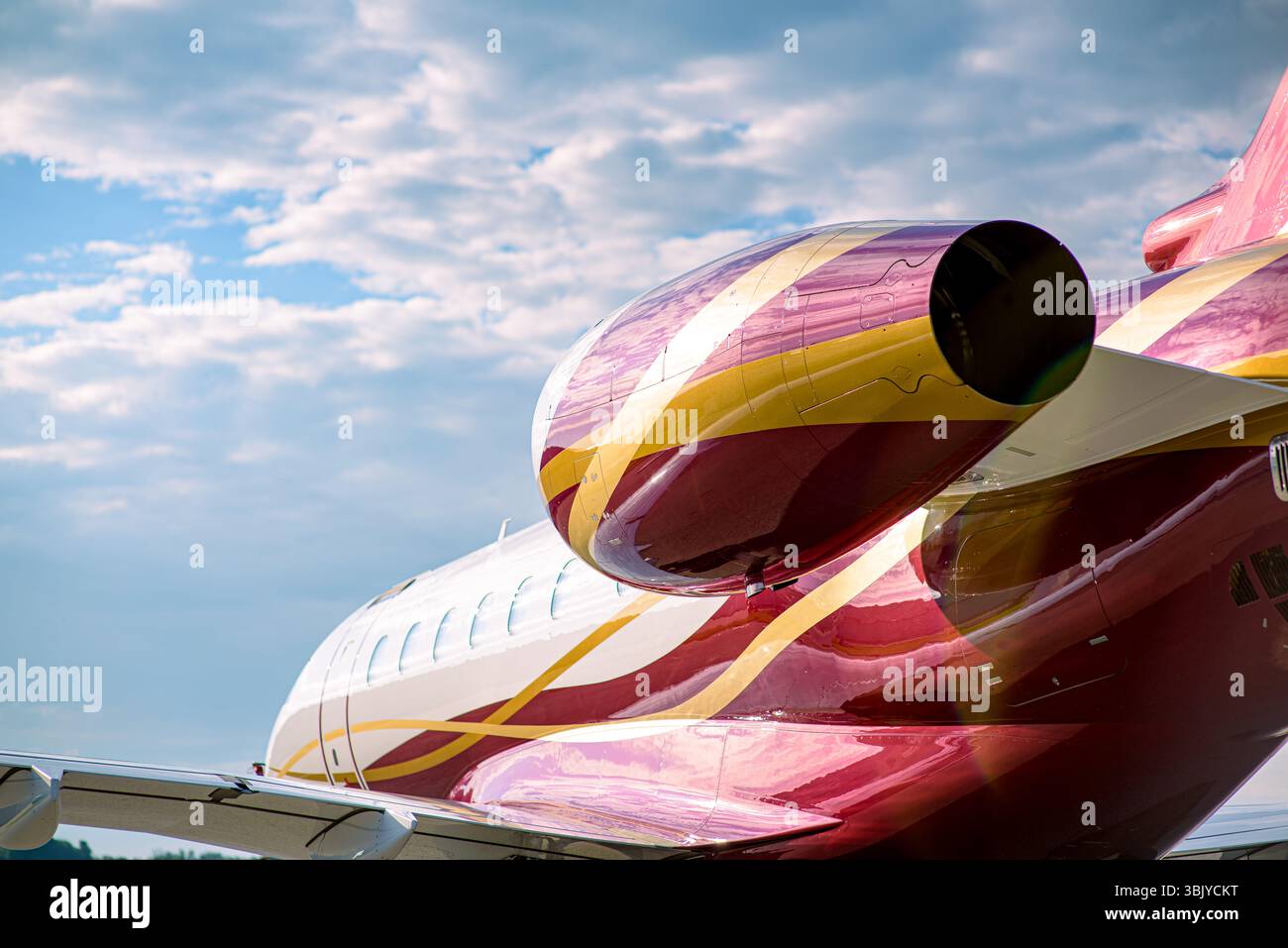 Business jet rosso e oro con navicella motore, finestrini cabina e sezione coda nel telaio. Fusoliera lucida che riflette la luce del sole sotto un cielo spettacolare. Foto Stock
