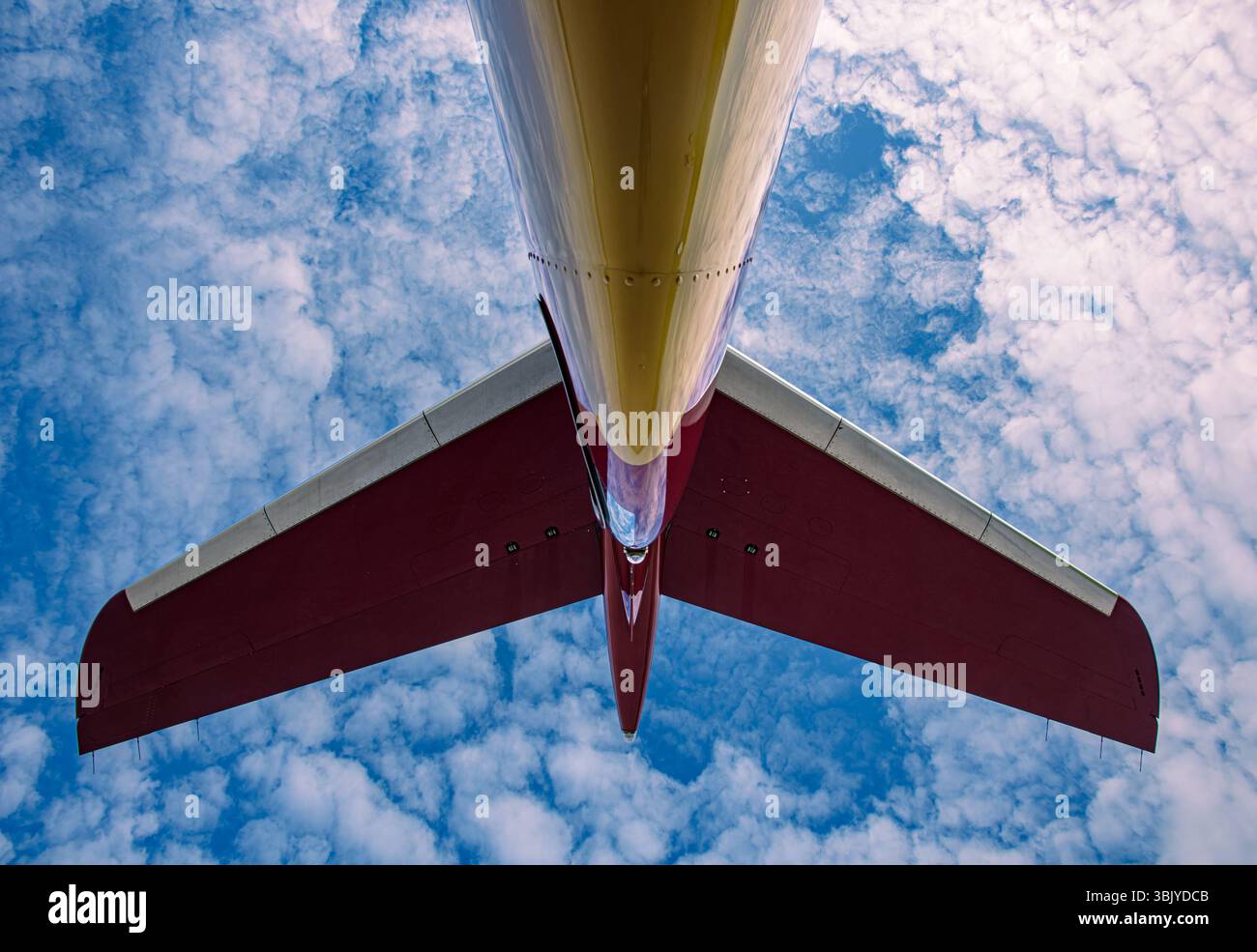 Vista dal basso verso l'alto di una sezione di coda del getto contro un cielo blu con nuvole. Fusoliera rossa e dorata con un design pulito dello stabilizzatore orizzontale. Foto Stock