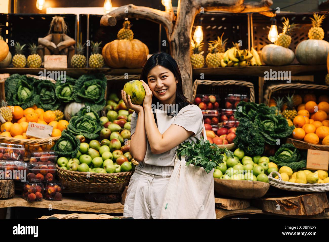 Donna allegra che tiene la frutta e guarda una macchina fotografica mentre si trova in piedi su un mercato locale all'aperto Foto Stock
